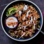Lucknowi biryani in a bowl topped with raita and a ramekin of raita on the side