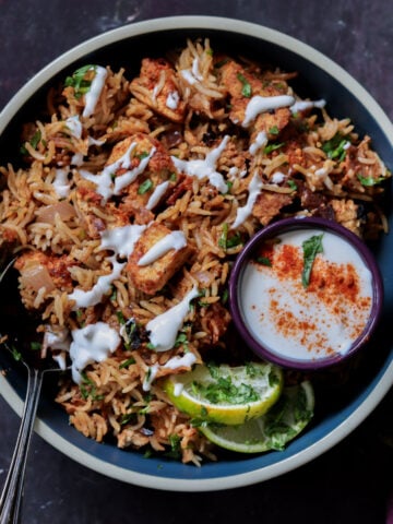 Lucknowi biryani in a bowl topped with raita and a ramekin of raita on the side