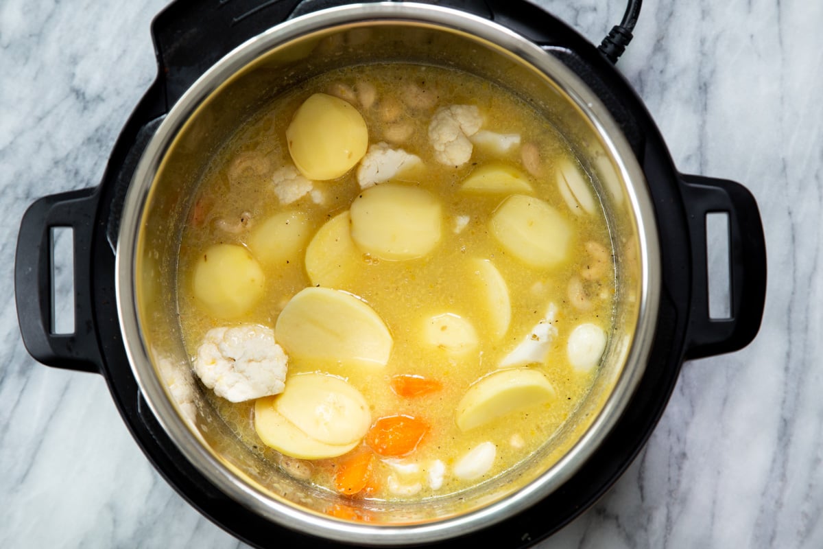 potato slices, cauliflower florets, nuts and carrots being added to an Instant pot filled with water and spices to make cheese sauce