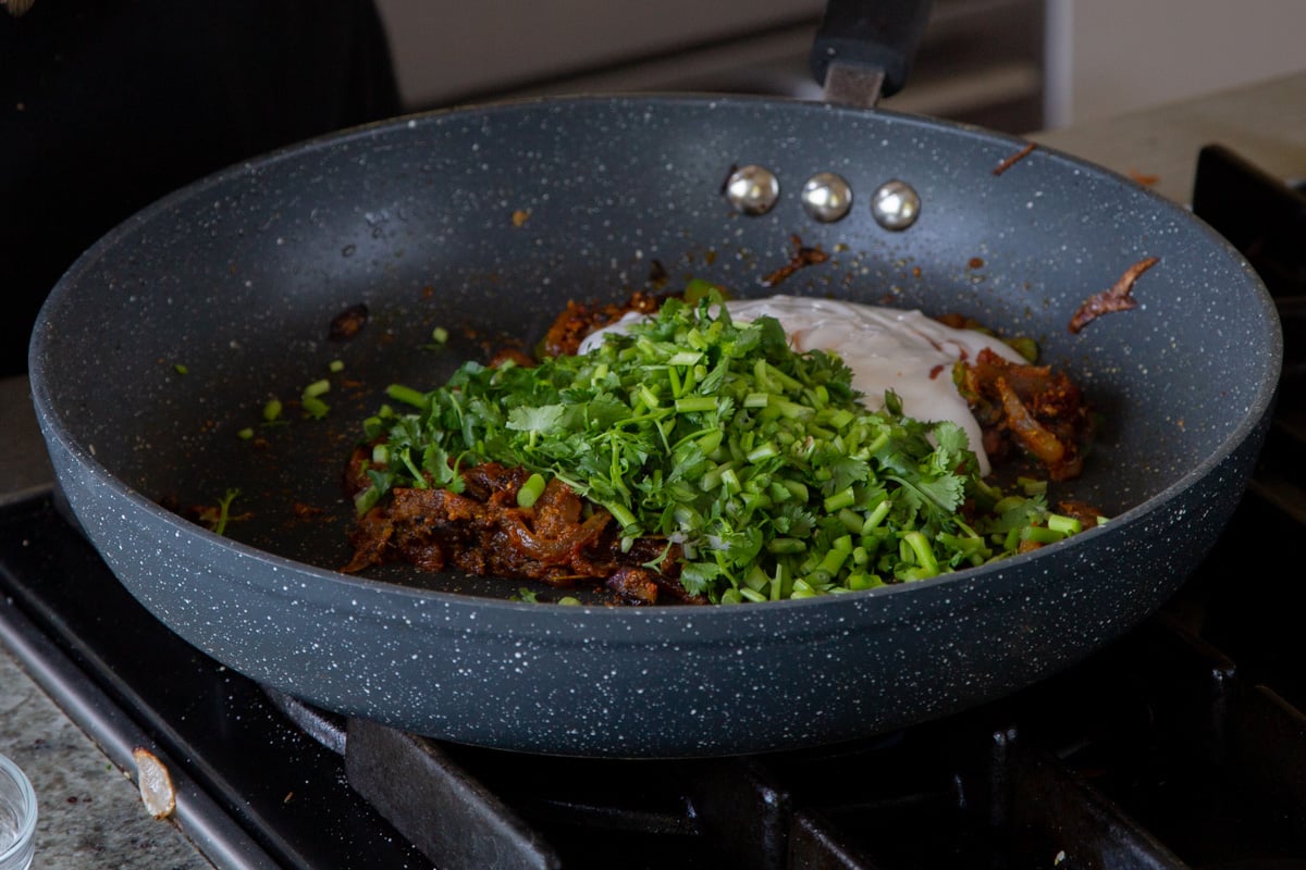 adding non-dairy yogurt and cilantro stems to the pan after mixing in the spices